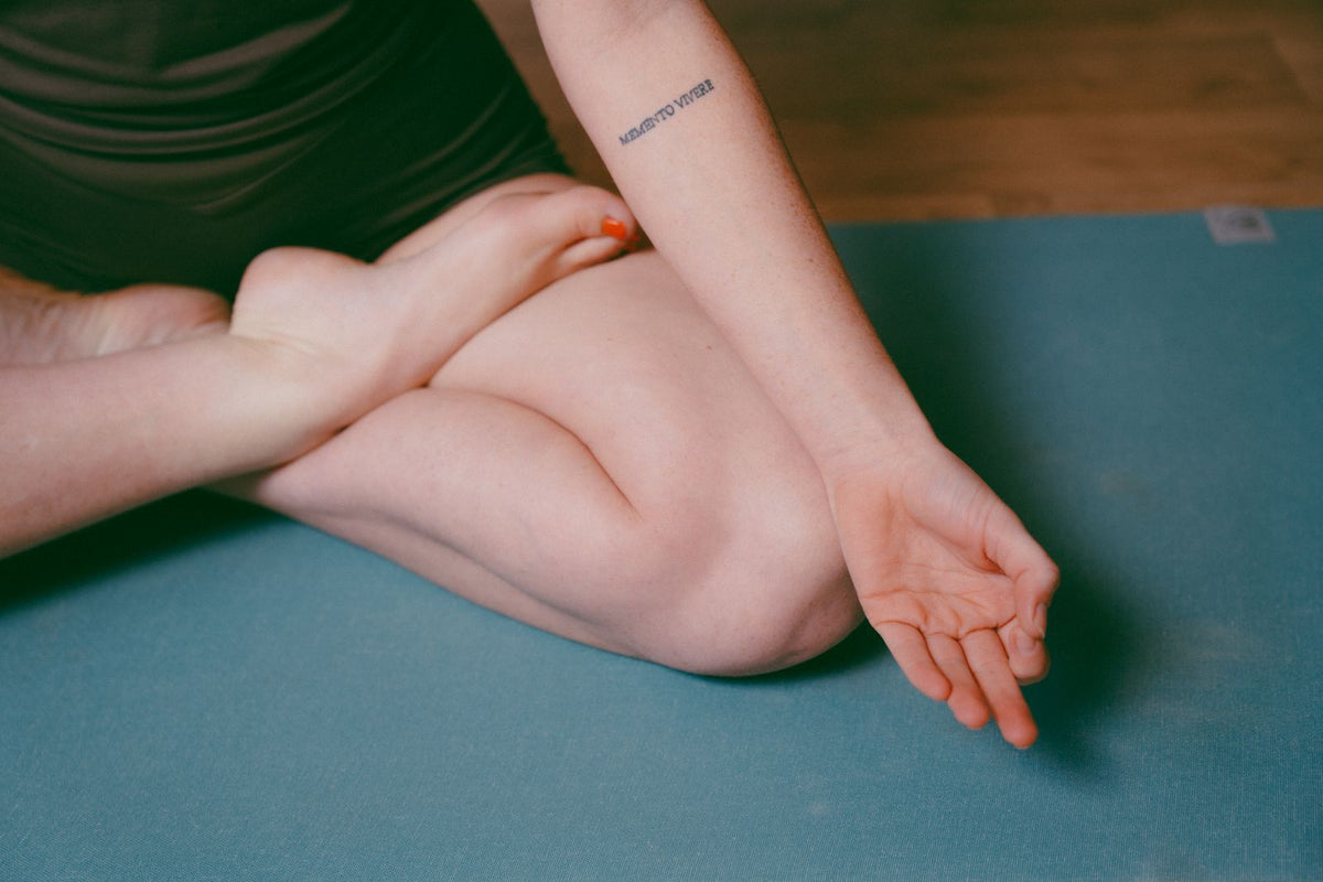 A close-up lifestyle shot of a person sitting cross-legged on a teal yoga mat, wearing a dark green athletic top. The person's crossed legs and one arm are clearly visible, with a small tattoo on the forearm.